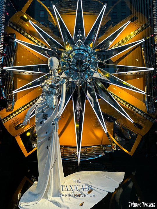 mannequin wearing a long white dress with the top of a yellow taxi cab blown up in the background as part of the 2024 Bergdorf Gooodman holiday window display in NYC