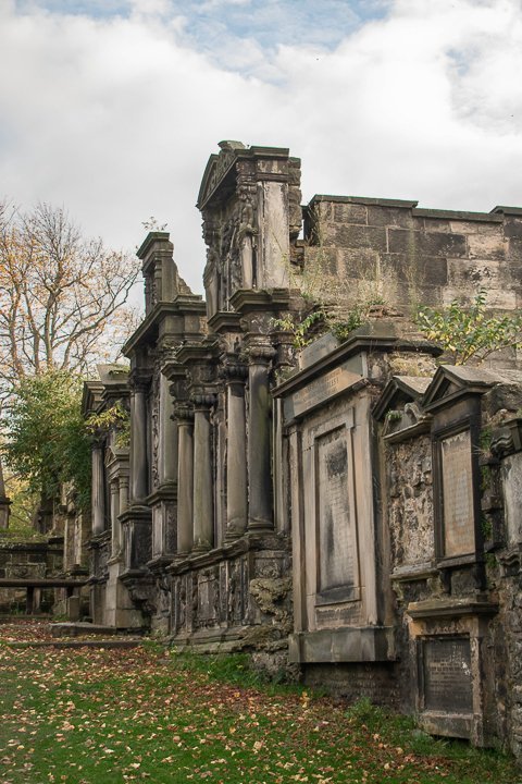 gray and brown stone graveyard structures with green grass, fall leaves against a cloudy sky in edinburgh scotland