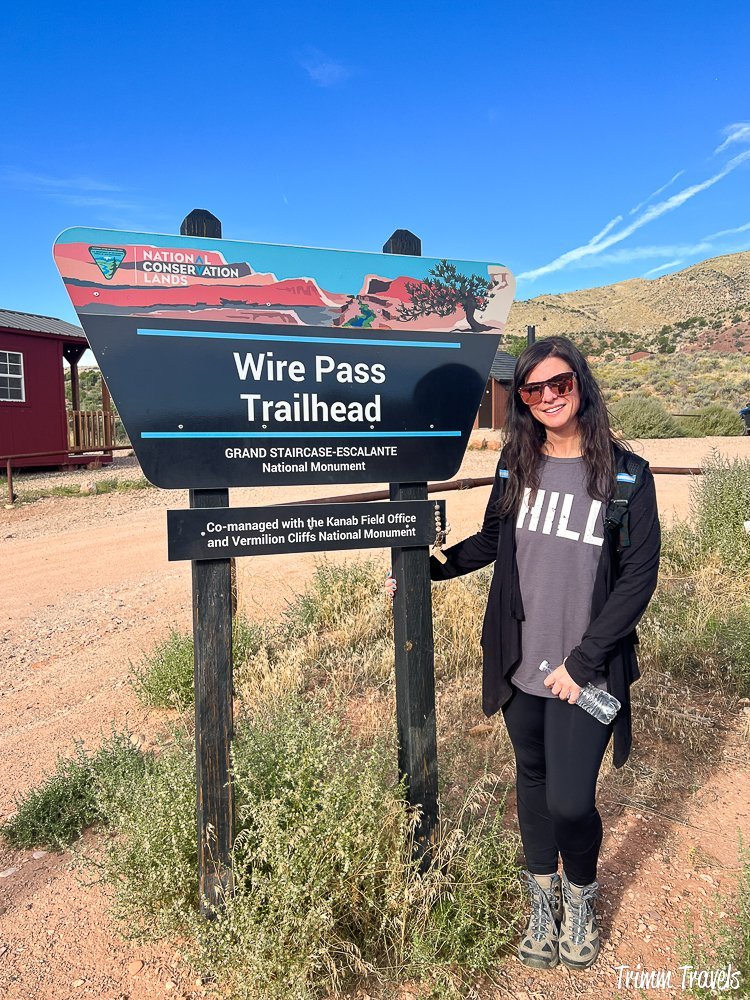 heather from trimm travels posing with the wire pass trailhead sign against a blue sky in utah