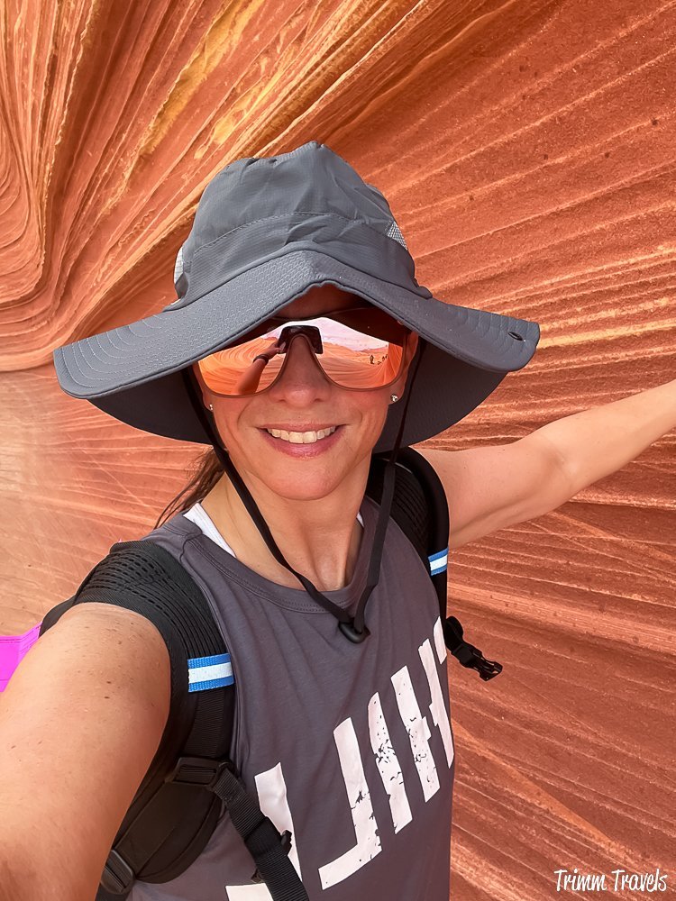 closeup of heather from trimm travels in a gray floppy sunhat and tank top up against a tall orange swirly sandstone formation of the wave in arizona