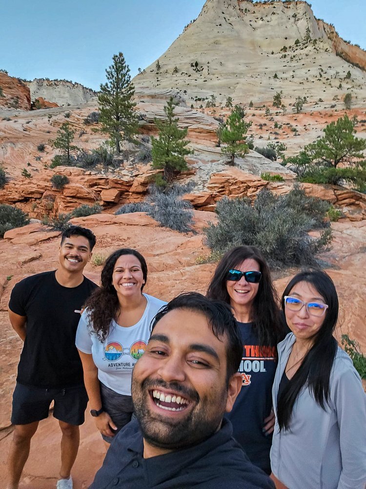 selfie of five of the six of us at zion national park with rocky sandstone formations in the background against a blue sky in utah