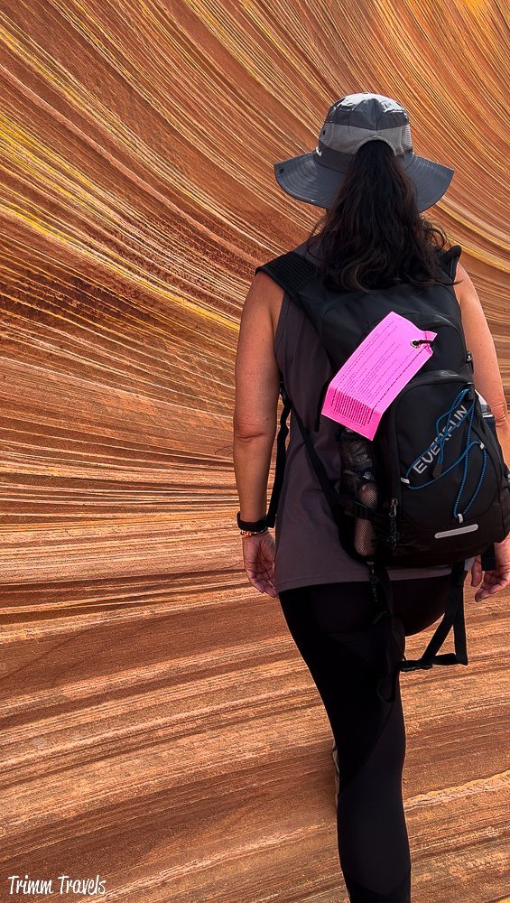 closeup of heather from trimm travels against the swirly sandstone formations of the wave in arizona and featuring the hot pink permit pinned to her black backpack