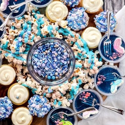overhead shot of a charcuterie board with blue jello cups, mini cupcakes, popcorn and blue and purple sprinkles against grey wood backdrop