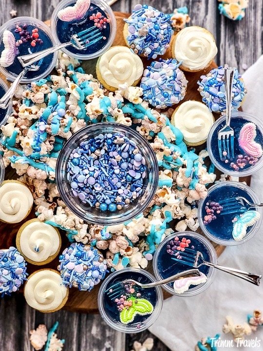 overhead shot of a charcuterie board with blue jello cups, mini cupcakes, popcorn and blue and purple sprinkles against grey wood backdrop