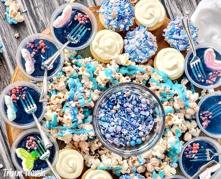 overhead shot of a charcuterie board with blue jello cups, mini cupcakes, popcorn and blue and purple sprinkles against grey wood backdrop