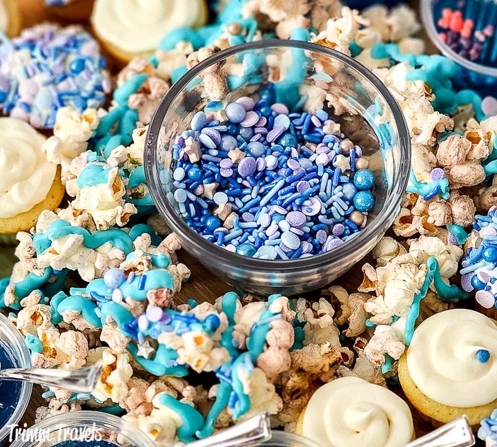 close up of a mini bowl of blue and purple sprinkles surrounded by white mini cupcakes and decorated popcorn