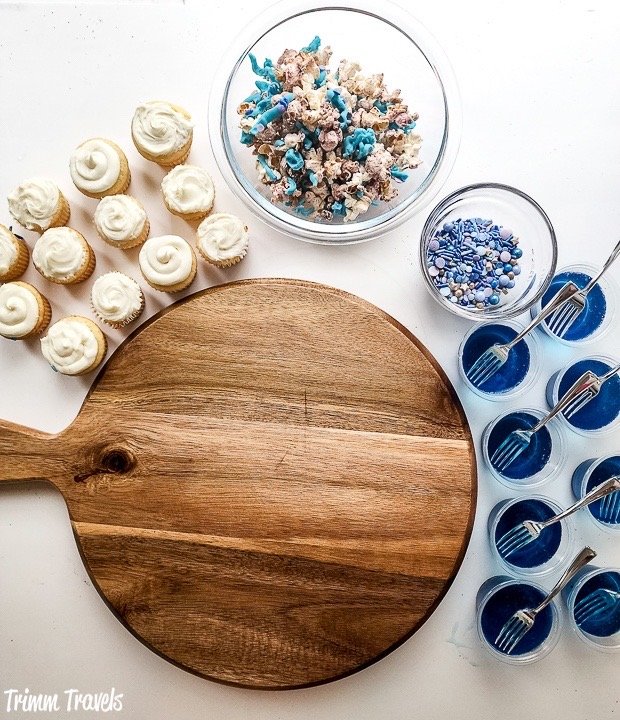 empty dark brown charcuterie board surrounded by mini white cupcakes, decorated popcorn, blue and purple sprinkles and blue jello cups against a white background