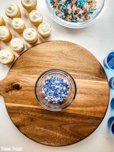 brown charcuterie board with a small bowl of purple and blue sprinkles surrounded around the board by mini white cupcakes and decorated popcorn