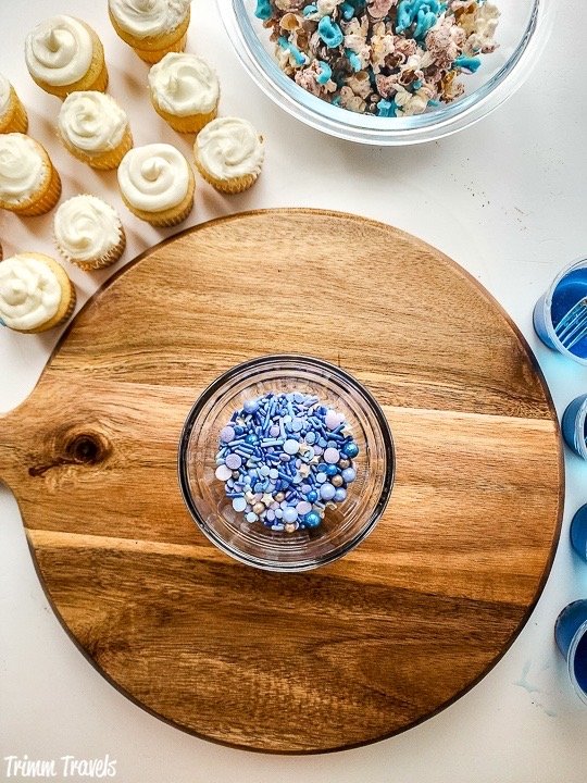 brown charcuterie board with a small bowl of purple and blue sprinkles surrounded around the board by mini white cupcakes and decorated popcorn