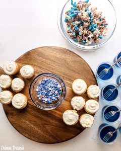 overhead shot of brown charcuterie board partially filled with mini cupcakes and blue and purple sprinkles surround by decorated popcorn and blue jello cups against white background