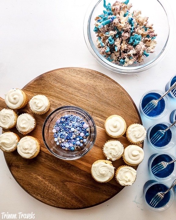 overhead shot of brown charcuterie board partially filled with mini cupcakes and blue and purple sprinkles surround by decorated popcorn and blue jello cups against white background
