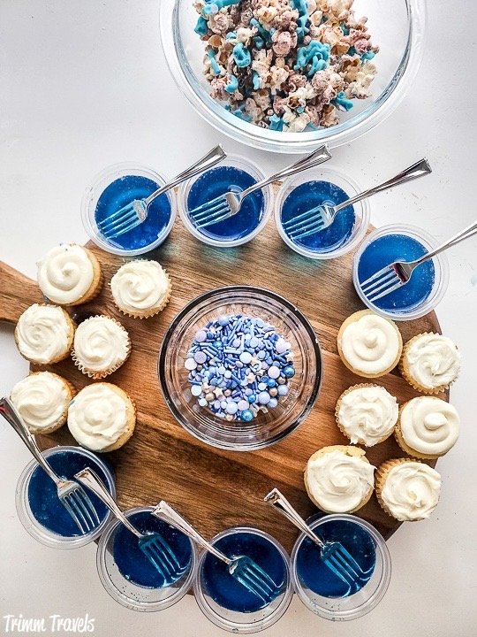 brown charcuterie board with mini white cupcakes, blue and purple sprinkles and blue jello cups with forks against a white background
