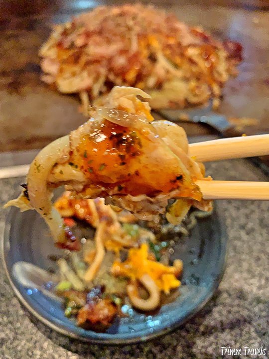 piece of okonomiyaki, a japanese pancake, on chopsticks above the rest of the pancake on the hibachi