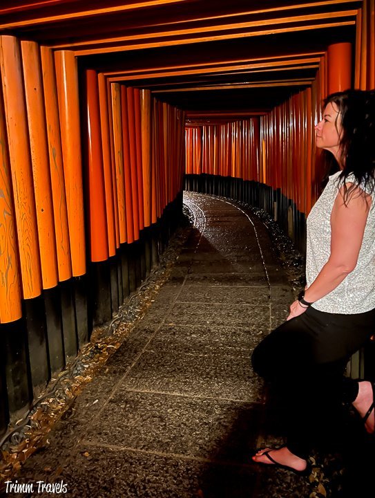 heather from trimm travels leaning up against the right side of the orange torii gates of fushimi inari at night
